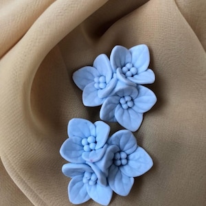 Op de afbeelding: Twee lichtblauwe bloemvormige versieringen. Elke bloem heeft vijf bloemblaadjes en een cluster van kleine, ronde details in het midden. De bloemen zijn geplaatst tegen een beige stoffen achtergrond.
