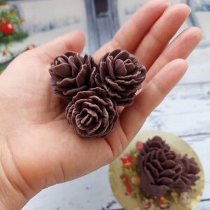 Könnte beinhalten: Drei dunkelbraune, rosenförmige Dekorationselemente. Das detaillierte Blumendesign ist sichtbar, mit mehreren Blütenblättern, die ein realistisches Aussehen erzeugen. Die Elemente werden in einer Hand gehalten, mit einem unscharfen Hintergrund.