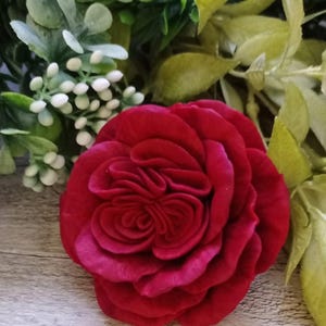 May include: A deep red, fabric rose with layered petals. The flower is the focal point, set against a backdrop of green and white foliage. The rose's texture and color create a striking visual contrast.