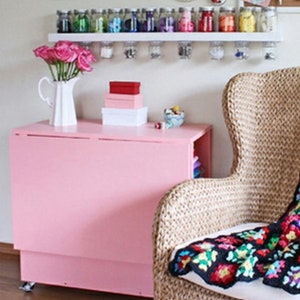 May include: A pink folding table with a white top and a wicker chair with a colorful crocheted throw blanket. The table has a shelf underneath and is decorated with a vase of pink flowers and a few boxes.