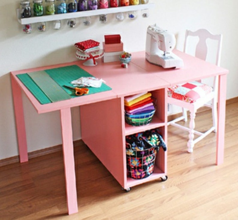 May include: A pink folding table with a sewing machine on top and storage shelves underneath. The table has a green cutting mat and a basket of colorful fabric scraps.