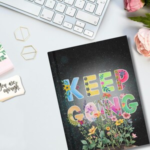 Puede incluir: Un cuaderno con las palabras "KEEP GOING" en letras coloridas, decorado con diseños florales. El fondo es un cielo oscuro y estrellado. Un teclado y artículos de papelería están en el fondo.