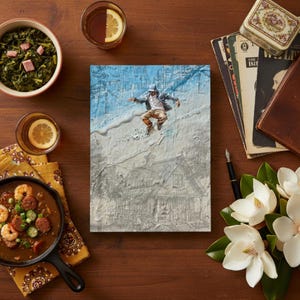 May include: A wooden table displays a canvas art piece with a person jumping, surrounded by food and drinks. A bowl of greens, a skillet of gumbo, and glasses with lemon slices are visible. Vintage books, a pen, and flowers complete the scene.