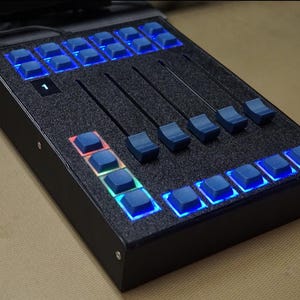 Puede incluir: Un panel de control electrónico negro con múltiples botones y deslizadores iluminados en azul. El panel tiene una superficie texturizada y una pequeña pantalla que muestra el número 1. Los botones y deslizadores están dispuestos en filas y columnas.