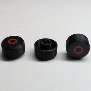 May include: Three black, cylindrical objects with a red ring in the centre. The objects appear to be 3D-printed and have a ridged texture. One object has a star-shaped interior.