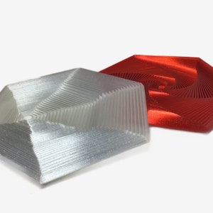 Pode incluir: Duas formas geométricas impressas em 3D, uma prateada e outra vermelha, com um padrão em espiral. As formas são hexagonais e têm um centro elevado.