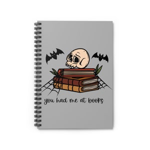 May include: A grey spiral-bound notebook featuring a stack of books, a skull, bats, and a spiderweb design. The text "you had me at books" is printed on the notebook.
