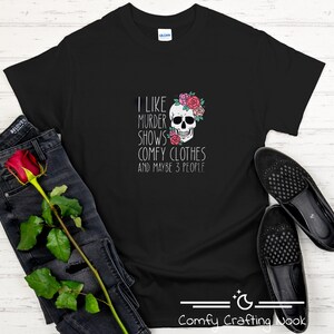 May include: Black t-shirt with a white graphic design featuring a skull with flowers and the text "I like murder shows, comfy clothes and maybe 3 people".