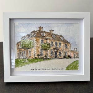 May include: A framed watercolour painting of a large stone manor house with a light-coloured driveway and green trees. The text at the bottom reads 26-28th June 2025 - Katie & Adam - Crucis Park, Cirencester. The artwork is enclosed in a white frame.