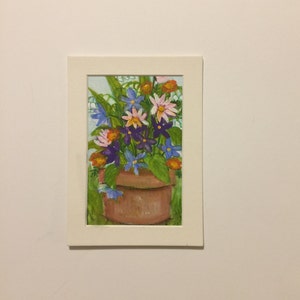 May include: A rectangular painting in a white mat frame depicting a terracotta pot filled with a vibrant bouquet of flowers. The arrangement features blue, purple, pink, and orange blossoms with green leaves, all rendered in visible brushstrokes.