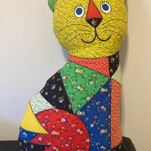 Könnte beinhalten: Ein handgefertigtes Kissen in Katzenform mit Patchwork-Design. Die Katze hat ein gelbes Gesicht mit blauen Augen, eine rote Nase und grüne Ohren. Der Körper besteht aus bunten Blumenstoff-Flicken.