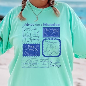 May include: A light teal t-shirt with the text "Advice from a Manatee" and manatee-themed illustrations in blue. The shirt features a relaxed fit and is worn by a person on a beach, with a straw hat and sunglasses.