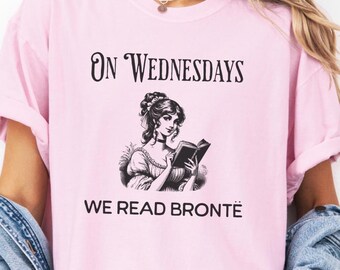 May include: A light pink t-shirt with the text "On Wednesdays We Read Brontë" in black. The shirt features a black and white illustration of a woman reading a book. The shirt is made of a soft material.