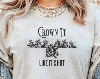 Może przedstawiać: Jasnoszara koszulka z napisem "Crown It" nad grafiką tańczących postaci i napisem "Like It's Hot" poniżej. Koszulka wykonana jest z miękkiego materiału.