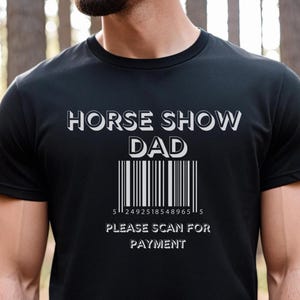 以下が含まれることがあります： 「HORSE SHOW DAD」の文字とバーコードがプリントされた黒いTシャツ。バーコードの下には「PLEASE SCAN FOR PAYMENT」の文字。クルーネックのTシャツです。