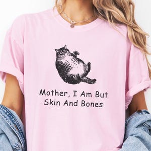 May include: A light pink t-shirt featuring a black illustration of a cat lying on its back. The text "Mother, I Am But Skin And Bones" is printed below the cat. The shirt has rolled-up sleeves.