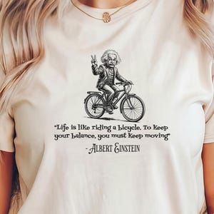 Op de afbeelding: Crèmekleurig T-shirt met een zwart-wit illustratie van Albert Einstein op een fiets. Het shirt bevat de quote "Het leven is als fietsen. Om je evenwicht te bewaren, moet je in beweging blijven" van Albert Einstein.