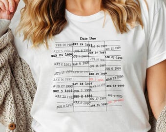 May include: A white t-shirt featuring a black and white graphic of a library card with the text "Date Due" and a list of dates. The dates are in a column format with the month, day, and year.