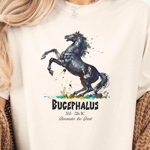 May include: Cream-colored t-shirt featuring a watercolor illustration of a rearing black horse with colorful accents. The text "BUCEPHALUS 355-326 BC Alexander the Great" is printed below the horse image.