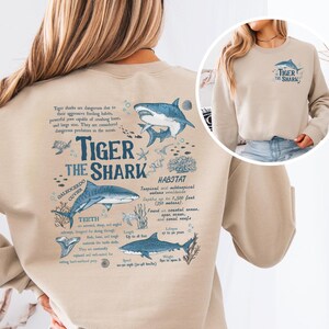 Könnte beinhalten: Beiges Sweatshirt mit detaillierter Tigerhai-Grafik und wissenschaftlichen Informationen. Der Text "TIGER THE SHARK" ist abgebildet. Eine kleinere Version des Designs befindet sich auf der oberen rechten Brust.