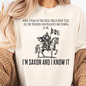以下が含まれることがあります： 「WHEN I WALK ON THE FIELD, THIS IS WHAT I SEE, ALL THE WENCHES AND PEASANTS ARE STARING AT ME. I'M SAXON AND I KNOW IT」のテキストがプリントされたクリーム色のTシャツ。馬に乗った騎士の白黒イラストが特徴です。