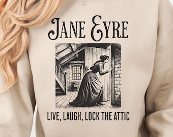 May include: A beige sweatshirt featuring the text "Jane Eyre" above a black and white illustration of a woman in a long dress. The text "Live, Laugh, Lock the Attic" is below the illustration. The sweatshirt has a crew neck.