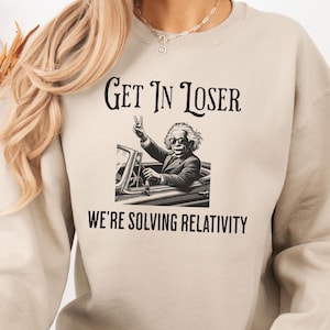 Puede incluir: Sudadera beige con el texto "Get In Loser" sobre una imagen de Albert Einstein en un coche, haciendo el signo de la paz. Debajo de la imagen está el texto "We're Solving Relativity".