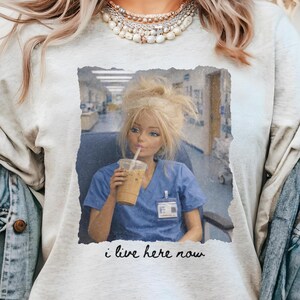 The Perfect Shirt for Nurses
The "I Live Here Now" nurse life shirt is a humorous ode to the demanding yet rewarding profession of nursing. Emblazoned with a comically frustrated doll sitting at a nurse's station clutching her essential iced coffee, this shirt captures the essence of long hours and dedication that nurses pour into their work. Ideal for new nurses, student nurses, or any medical professional, this funny nurse shirt makes for a thoughtful and amusing gift.