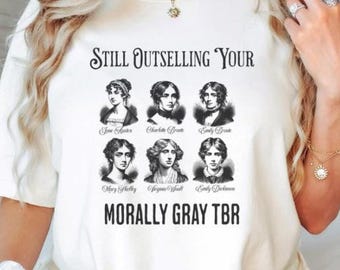 May include: Cream-colored t-shirt featuring the text "Still Outselling Your" above portraits of six female authors. Below the portraits, the text reads "Morally Gray TBR". The shirt is a casual, unisex style.