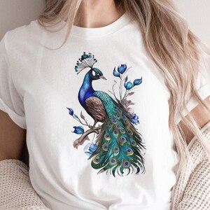May include: White t-shirt with a blue and green peacock design. The peacock is perched on a branch with blue flowers.