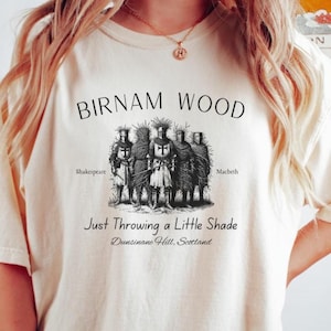 Puede incluir: Camiseta color crema con el texto "BIRNAM WOOD" sobre una ilustración en blanco y negro de figuras. Debajo, el texto dice "Just Throwing a Little Shade" y "Dunsinane Hill, Scotland."