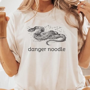 Pode incluir: Camiseta creme com uma ilustração em preto e branco de uma cobra e a frase "danger noodle" abaixo. A camiseta é de estilo casual, adequada para uso diário.
