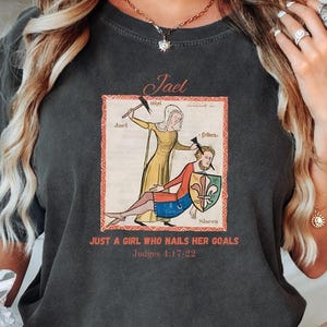 May include: Dark gray t-shirt featuring a medieval-style illustration of a woman holding a hammer over a man. The text reads "Jael" above the image and "JUST A GIRL WHO NAILS HER GOALS" below, with a Bible verse reference.