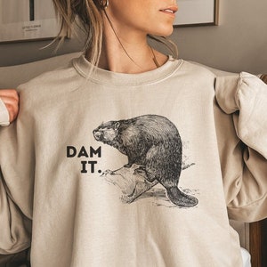May include: Beige sweatshirt with a black and white drawing of a beaver on a log. The text "DAM IT." is printed in black. The sweatshirt has a crew neck and long sleeves. The image is a humorous design.