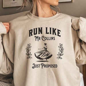 Puede incluir: Una sudadera beige con un estampado gráfico negro que dice "Run Like Mr. Collins Just Proposed". El gráfico presenta una silueta negra de una mujer con un vestido corriendo.