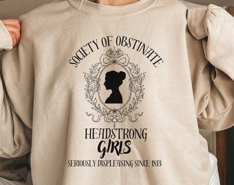 May include: Beige sweatshirt with a black graphic print that reads "Society of Obstinate Headstrong Girls Seriously Displeasing Since 1813". The graphic features a silhouette of a woman's head with a bun and a floral wreath.