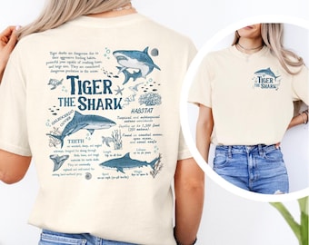 May include: Cream-colored t-shirt featuring a detailed graphic of a tiger shark with informational text. The design includes illustrations of sharks, coral, and text about the shark's habitat and characteristics. The shirt also has a smaller shark graphic on the front.