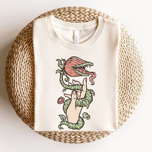 May include: A white t-shirt with a graphic of a hand being wrapped by a carnivorous plant with a red flower and green leaves.