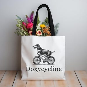 May include: White canvas tote bag with black handles. The bag features a black and white illustration of a dachshund riding a bicycle. The text "Doxycycline" is printed in black below the illustration.
