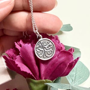 May include: A silver-toned circular pendant necklace featuring embossed Arabic calligraphy on its face. The pendant hangs from a delicate silver-toned chain, showcasing a metallic finish with dark recessed details that highlight the intricate script.