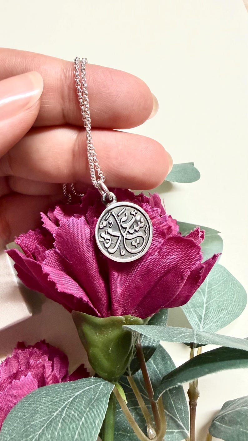 May include: A silver-toned circular pendant necklace featuring embossed Arabic calligraphy on its face. The pendant hangs from a delicate silver-toned chain, showcasing a metallic finish with dark recessed details that highlight the intricate script.