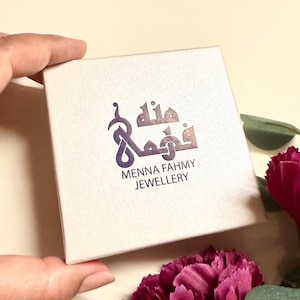 May include: A square, off-white jewelry box featuring a purple logo with Arabic calligraphy and the text "MENNA FAHMY JEWELLERY" printed in a dark font on the center of the lid. The box has a smooth, matte finish and a clean, simple design.