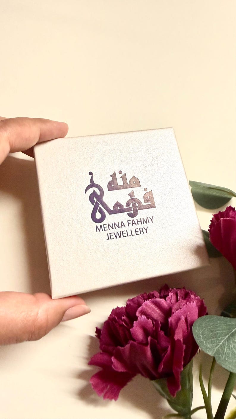 May include: A square, off-white jewelry box featuring a purple logo with Arabic calligraphy and the text "MENNA FAHMY JEWELLERY" printed in a dark font on the center of the lid. The box has a smooth, matte finish and a clean, simple design.