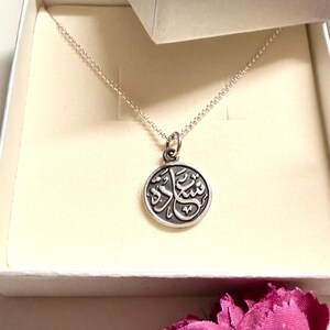 May include: A silver-toned circular pendant necklace featuring Arabic calligraphy, presented in a white jewelry box with the text Menna Fahmy Jewellery printed on the lid. The pendant hangs from a delicate silver-toned chain inside the box.