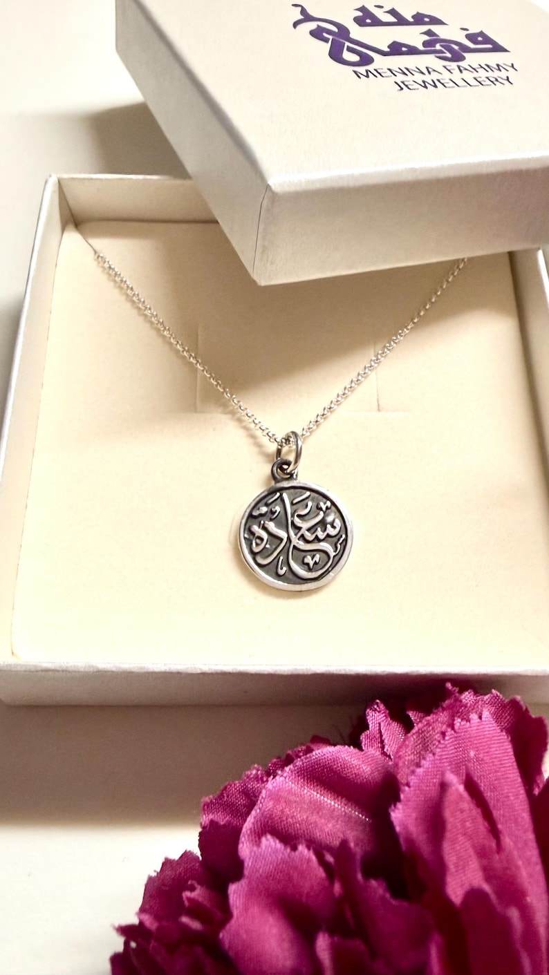 May include: A silver-toned circular pendant necklace featuring Arabic calligraphy, presented in a white jewelry box with the text Menna Fahmy Jewellery printed on the lid. The pendant hangs from a delicate silver-toned chain inside the box.