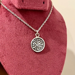 May include: A silver-toned necklace featuring a circular pendant with a dark, recessed background and raised silver Arabic calligraphy. The pendant hangs from a delicate silver-toned chain with small, uniform links, creating a simple and clean design.