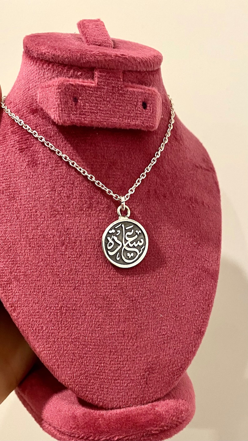 May include: A silver-toned necklace featuring a circular pendant with a dark, recessed background and raised silver Arabic calligraphy. The pendant hangs from a delicate silver-toned chain with small, uniform links, creating a simple and clean design.