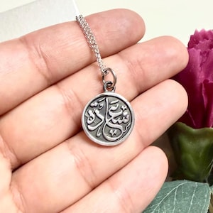 May include: A circular silver pendant on a delicate silver chain. The pendant features a raised, dark-oxidized Arabic script inscription in the center, set against a smooth, polished silver background with a defined circular border.