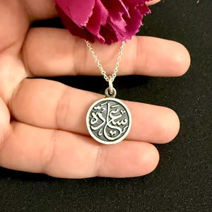 May include: A circular silver pendant on a delicate silver chain, featuring an engraved Arabic script design in a dark recessed background. The pendant is positioned below a vibrant magenta flower, showcasing a polished metallic finish.