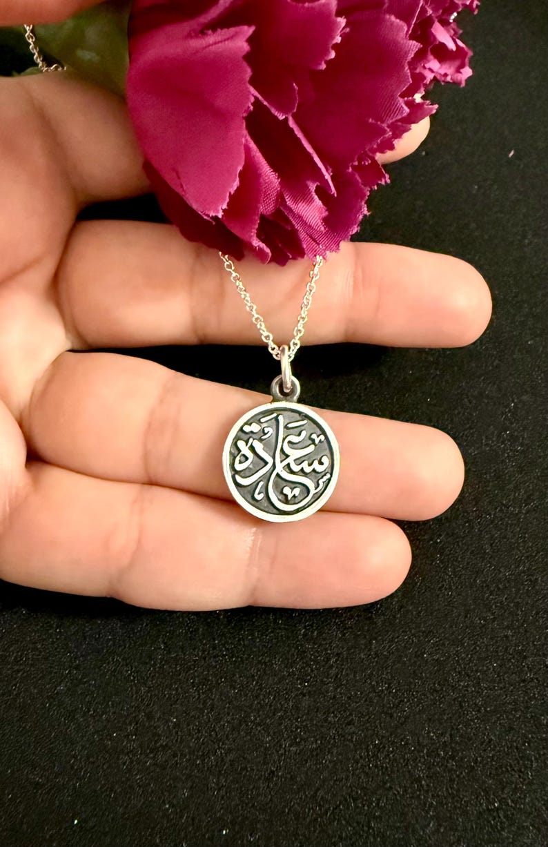 May include: A circular silver pendant on a delicate silver chain, featuring an engraved Arabic script design in a dark recessed background. The pendant is positioned below a vibrant magenta flower, showcasing a polished metallic finish.